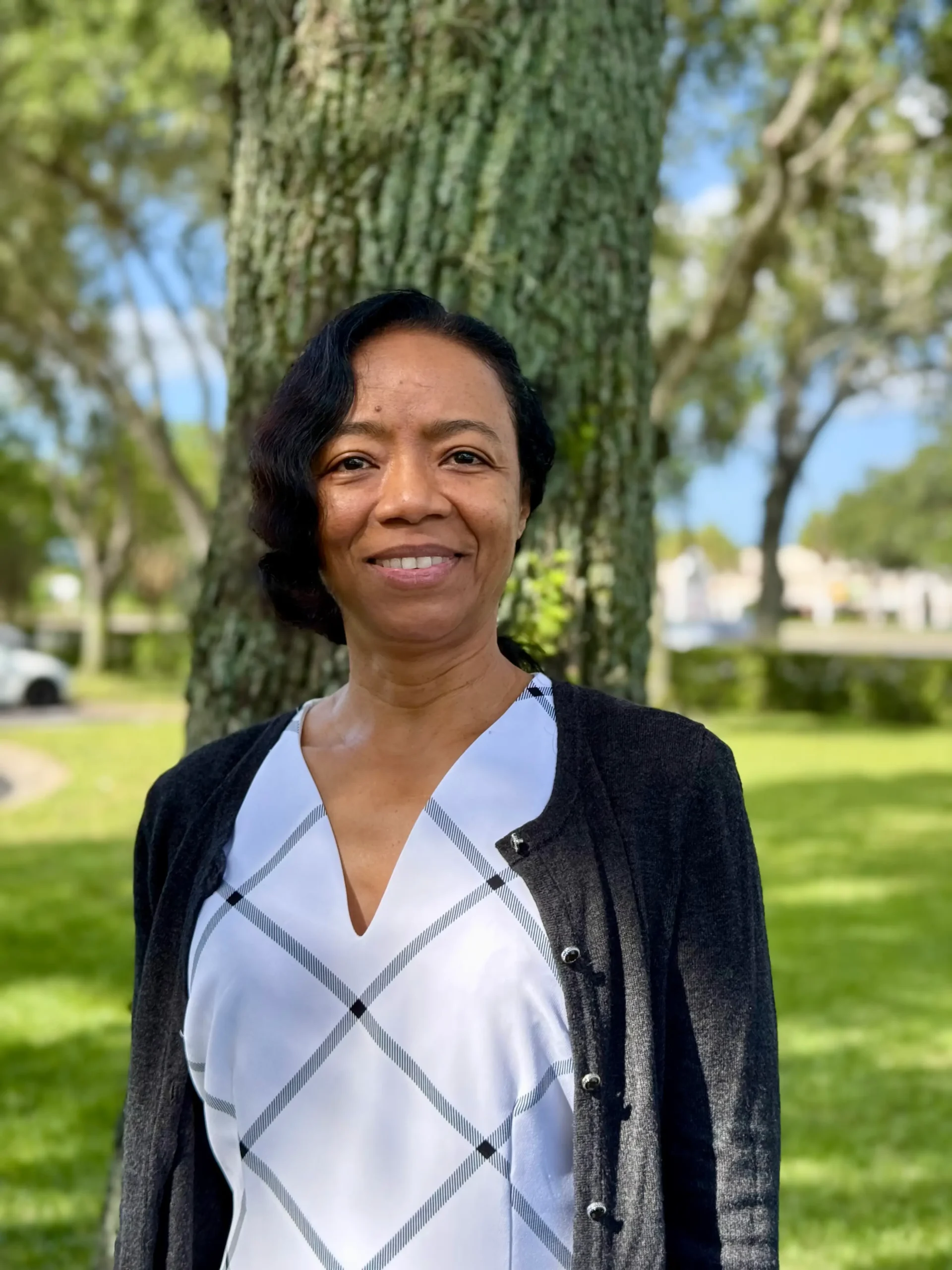 Portrait of a smiling woman standing outdoors in a park with a tree trunk behind her. In focus from the chest up, wearing a white plaid blouse and dark cardigan.