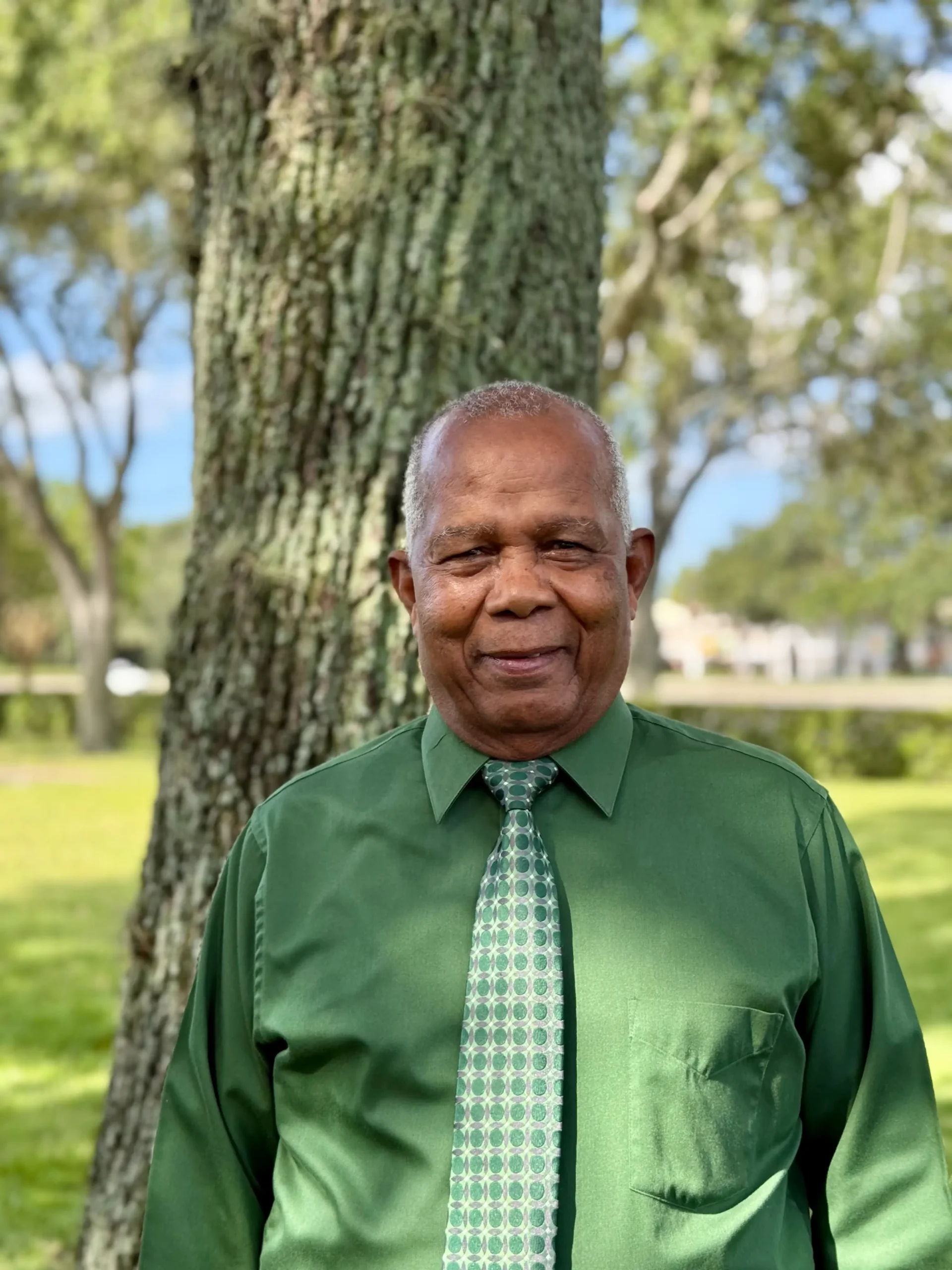 Smiling older man in a green shirt and patterned tie stands outdoors near a tree in a park setting.