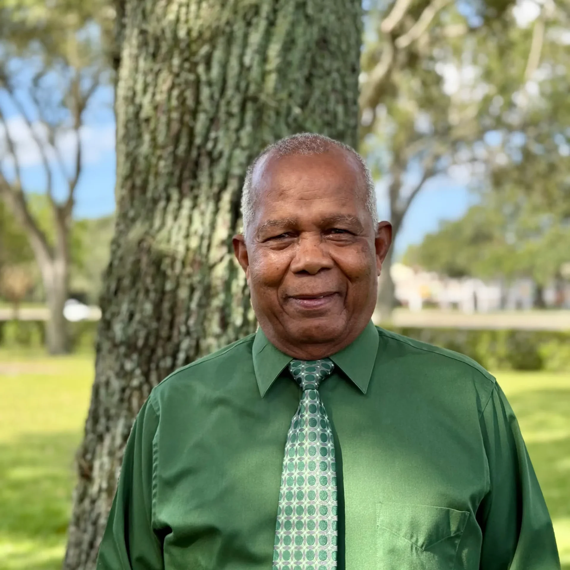 Smiling older man in a green shirt and patterned tie stands outdoors near a tree in a park setting.